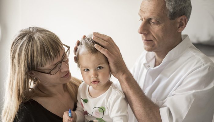 Osteopatas švelniai laiko kūdikio galvytę mamai laikant vaiką ant rankų